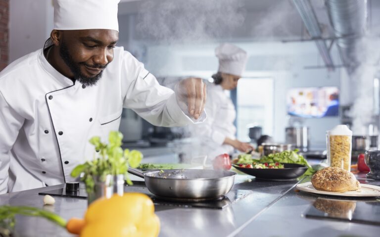 In a kitchen, black chef adding fresh herbs to complete a gourmet dish, balancing flavors with rosemary and other condiments. Kitchen counter stocked with vegetables and food prep essentials.